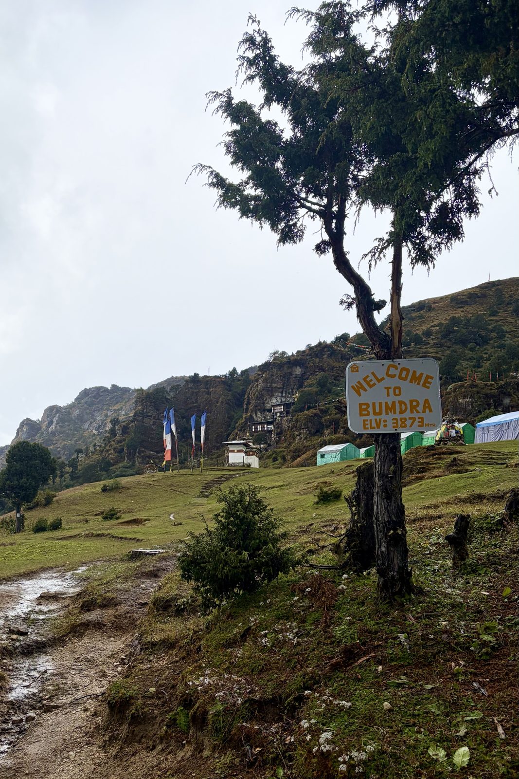 Een bordje aan een boom met de tekst Welcome to Bumdra, Elv: 3873 mtr, met daarachter een grazige helling, hoge gebedsvlaggen, een chorten, groene tenten en een klooster tegen een rotswand, onder een grijze lucht.