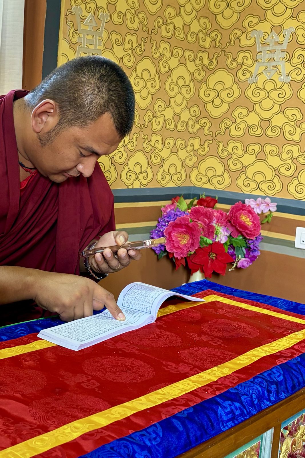 Een monnik in een rode pij buigt zich over een opengeslagen boekje met tabellen in Tibetaans schrift op een rood-blauw brokaatkleed en kijkt tegelijk op zijn gsm, met een geel beschilderde muur en een vaas bloemen erachter.