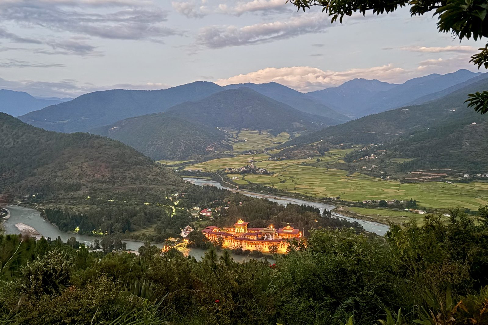 Een weids schemerzicht vanaf een helling: de Punakha Dzong gloeit goud van de lichtjes waar twee rivieren samenkomen, met rijstvelden en boerderijen die zich verder de vallei in uitstrekken, en gelaagde blauwe bergen onder een zachte avondhemel.