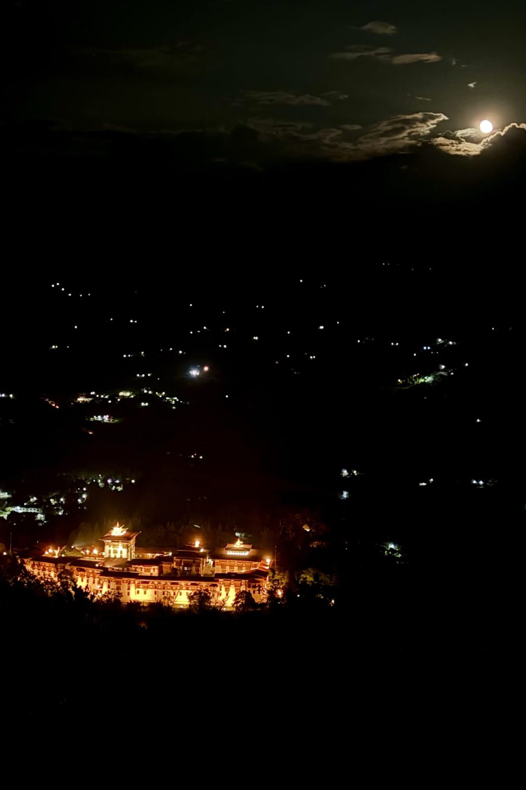 Een nachtzicht naar beneden in een donkere vallei: een grote dzong in warm goud verlicht onderaan, verspreide lichtjes van huizen op de hellingen, en een heldere maan die rechtsboven door een dunne wolkenlaag breekt.