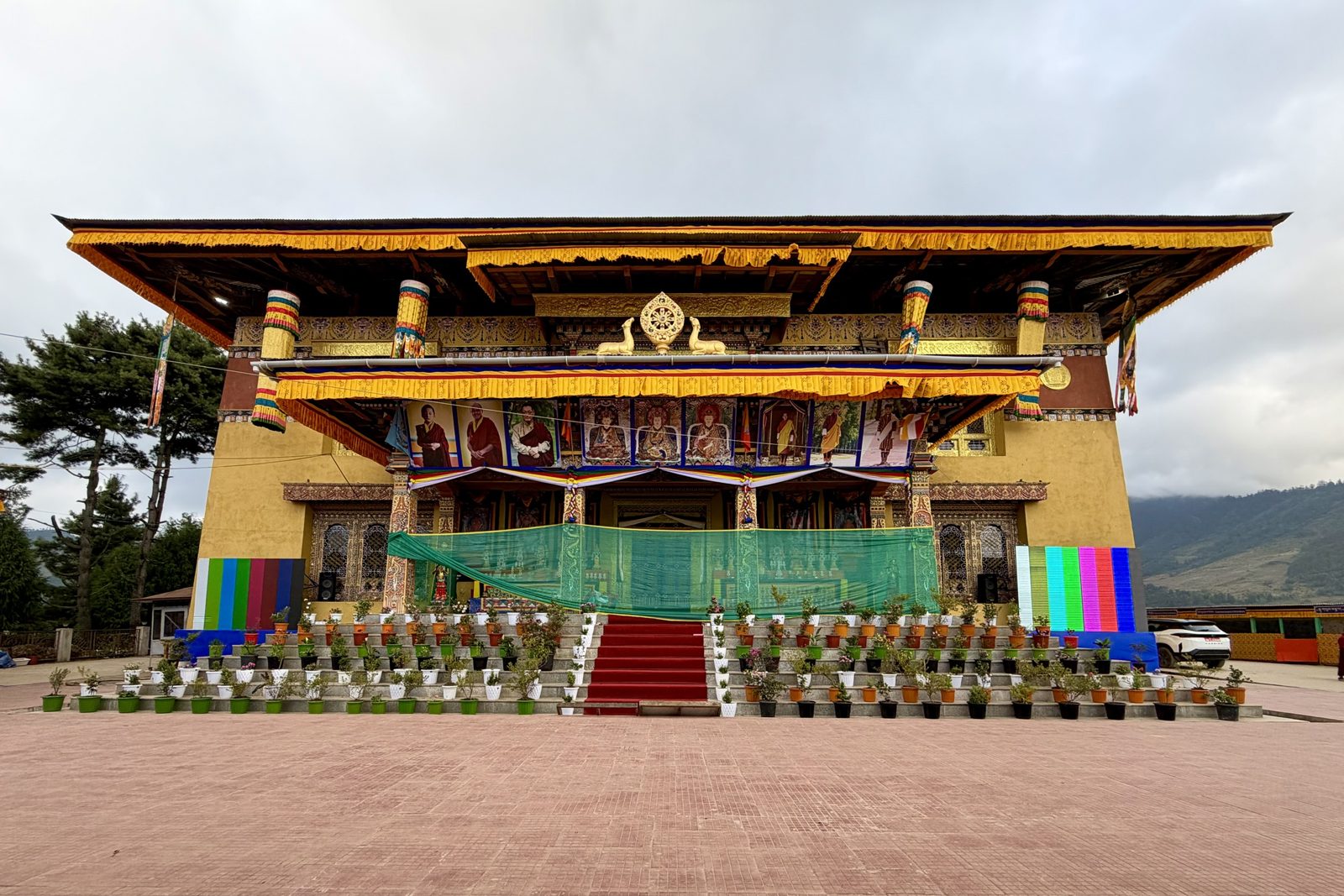 De gevel van een gele tempel met gouden dakranden en een dharmawiel met twee herten, een rij grote portretten van lama&rsquo;s en koningen, een groen net voor de veranda, beeldschermen aan weerszijden en trappen vol bloempotten met een rode loper.