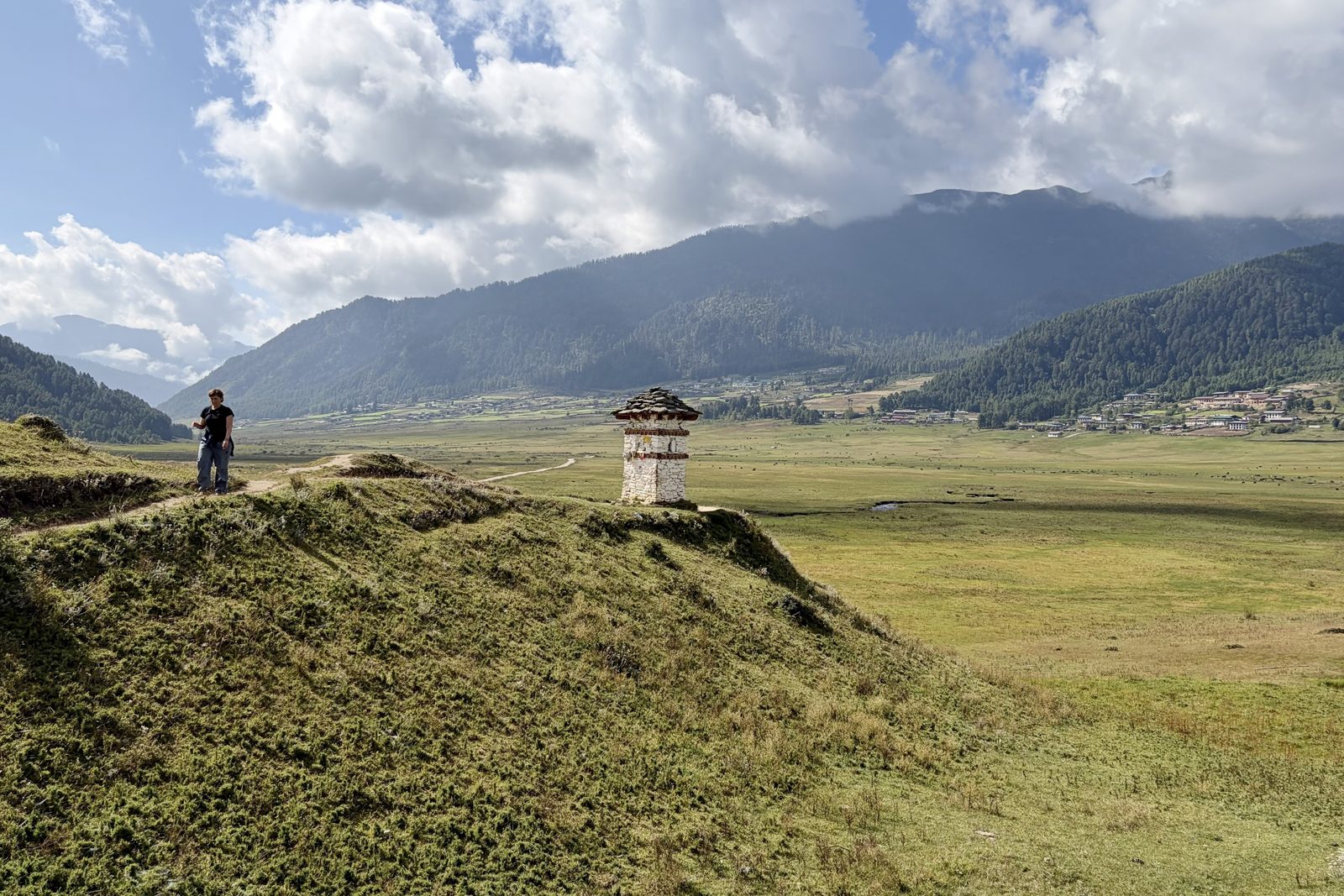 Iemand van ons op een grazige heuvelrug naast een klein wit gemetseld chorten-huisje met een dak van leisteen, met daarachter de brede vlakke vallei, dorpjes aan de bosrand en bergen in de wolken.