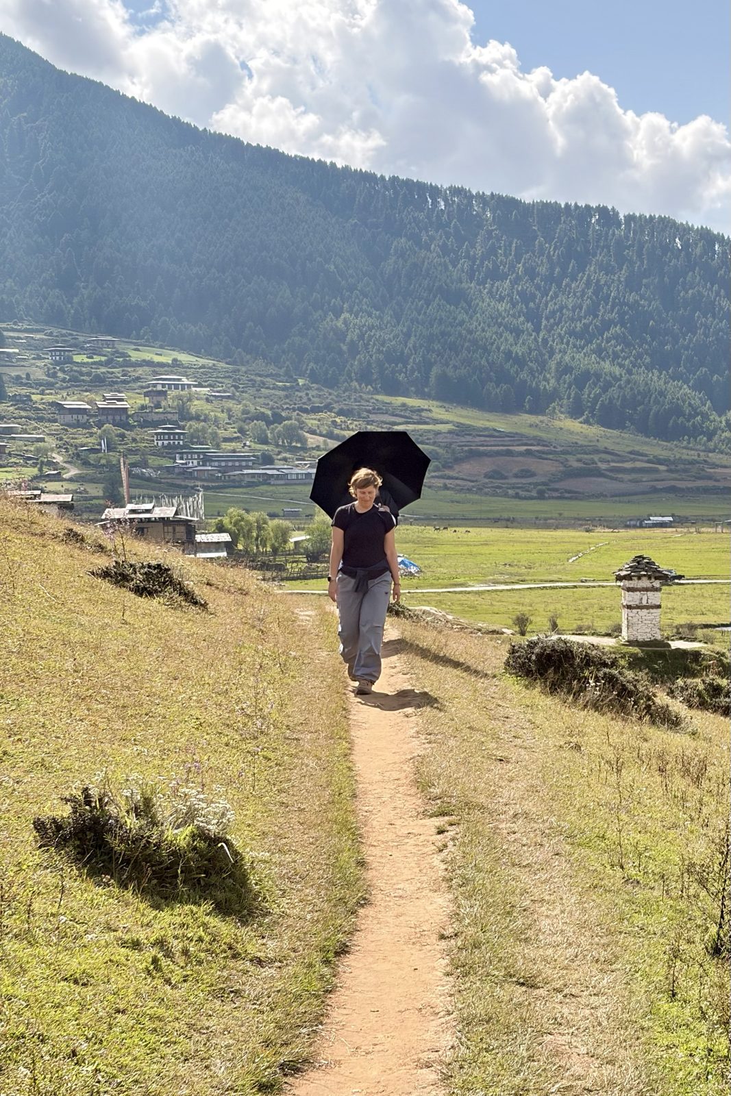 Iemand van ons stapt over een smal zandpad op een grazige helling, met een zwarte paraplu tegen de zon, met daarachter een dorp, gebedsvlaggen en een beboste berg.