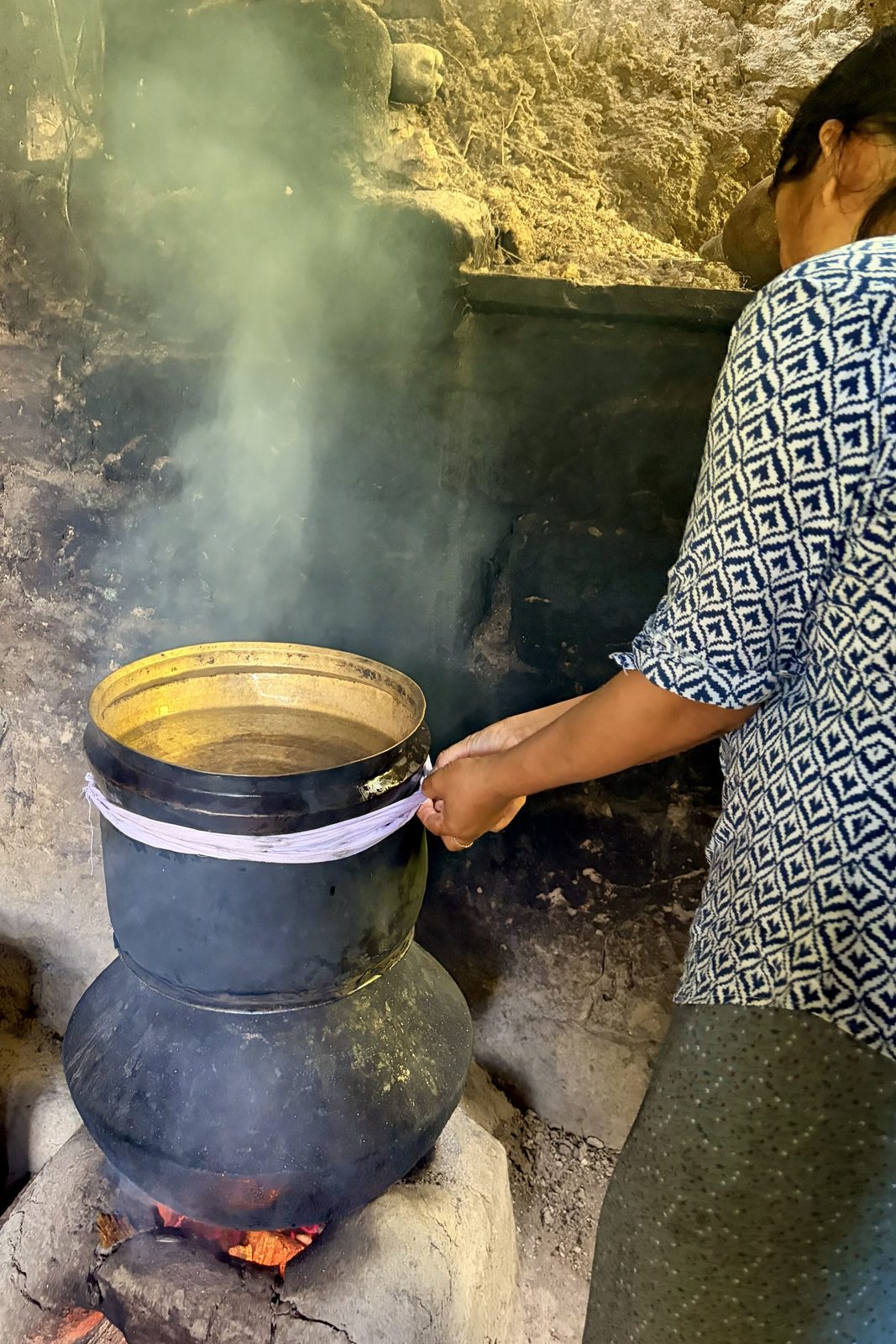 Een vrouw die een grote zwarte metalen distilleerketel boven een houtvuur verzorgt, in een door rook zwartgeblakerde keuken, met opstijgende stoom en een witte doek rond de naad tussen de onderste pot en de bovenste kom gewikkeld.