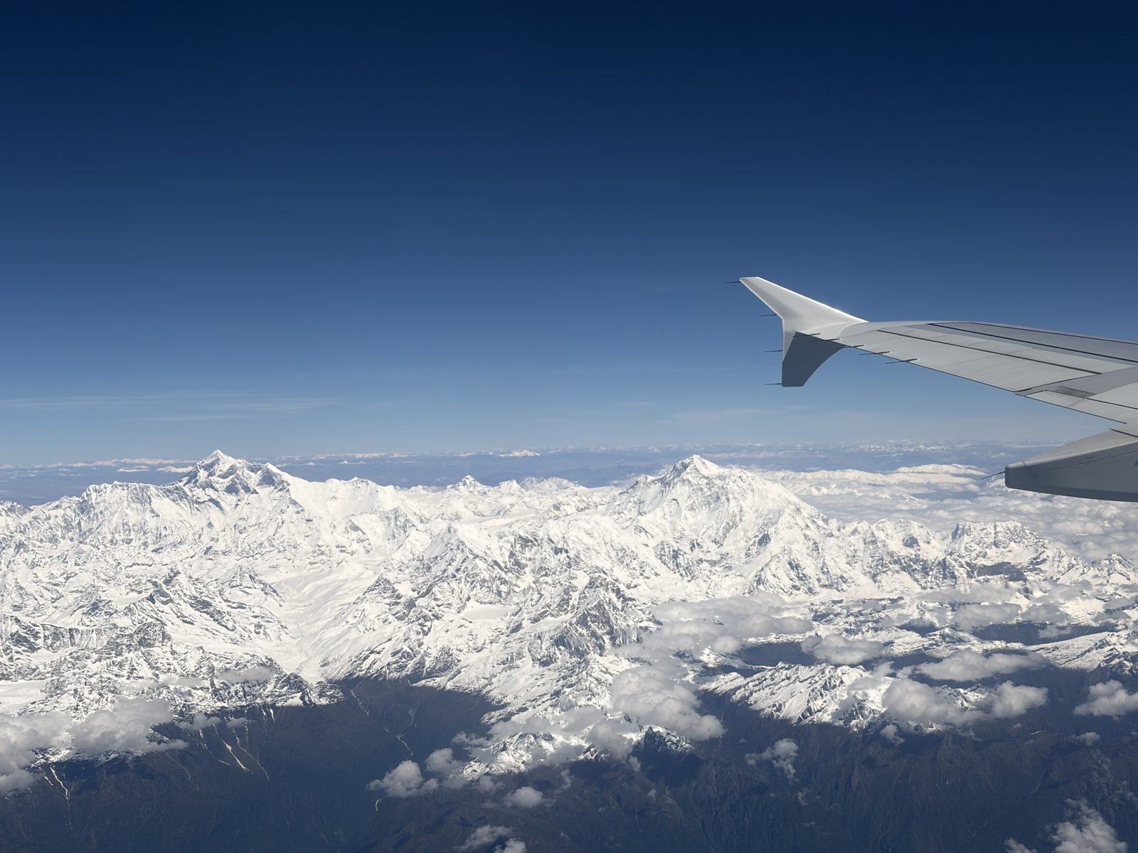 Besneeuwde Himalaya-keten onder een diepblauwe hemel, gezien door een vliegtuigraam met de vleugeltip rechts; twee hoge toppen steken boven de rest uit, met wolken die over de donkere valleien eronder drijven.