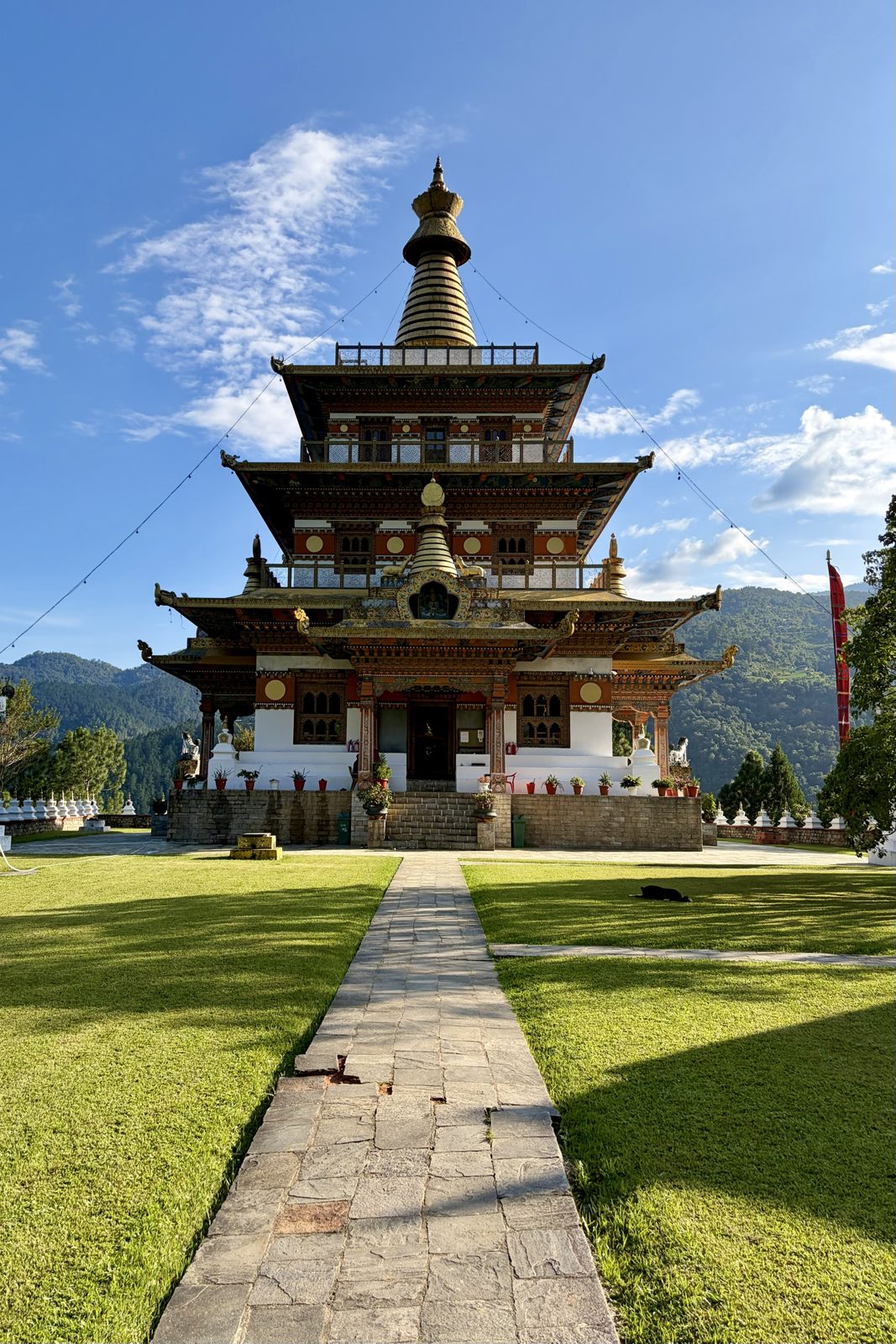 Een hoge tempel met drie verdiepingen, vergulde daken en een gouden spits, recht van voren gezien aan het eind van een stenen pad tussen twee zonnige gazons, met beboste heuvels erachter.