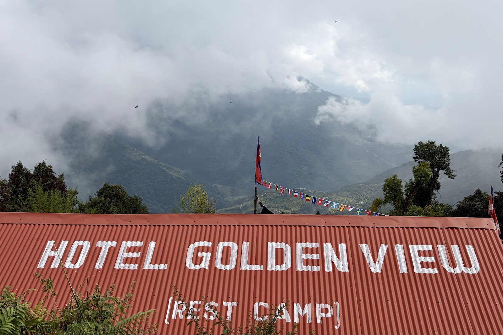 Een rood golfplaten dak met in witte letters Hotel Golden View Rest Camp, met daarachter wolken over beboste bergen.