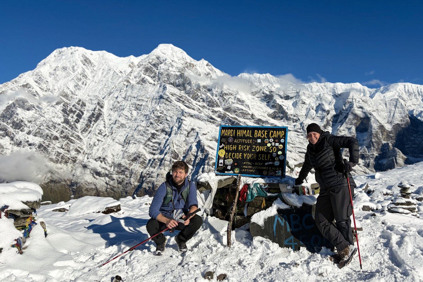 Twee mensen bij een bord van Mardi Himal Base Camp in de sneeuw, met een enorme besneeuwde bergwand en een blauwe lucht erachter.