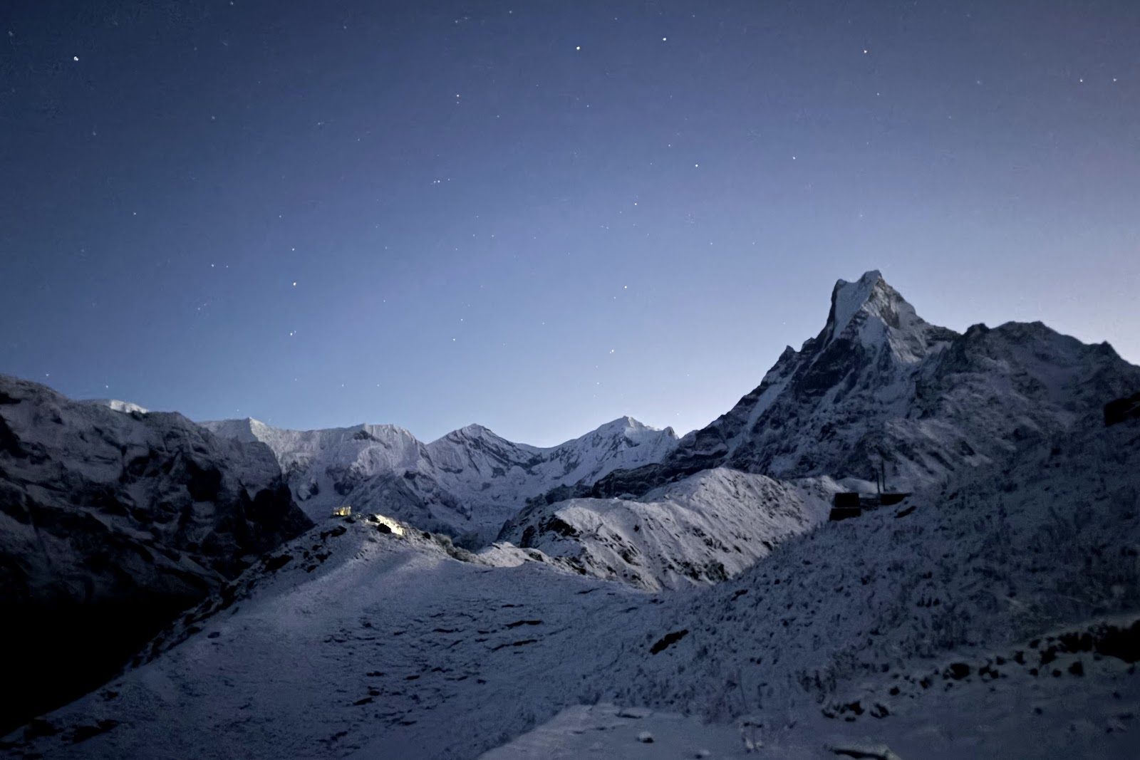 De spitse top van de Machhapuchhre en andere besneeuwde bergen onder een sterrenhemel in het eerste ochtendlicht.
