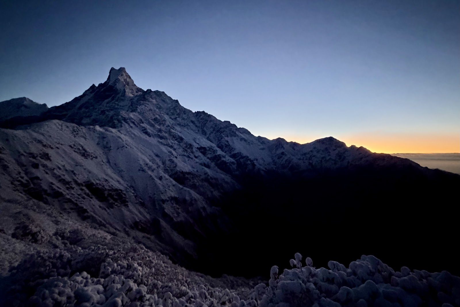 Het donkere silhouet van de Machhapuchhre tegen een oranje gloed aan de horizon bij zonsopgang.