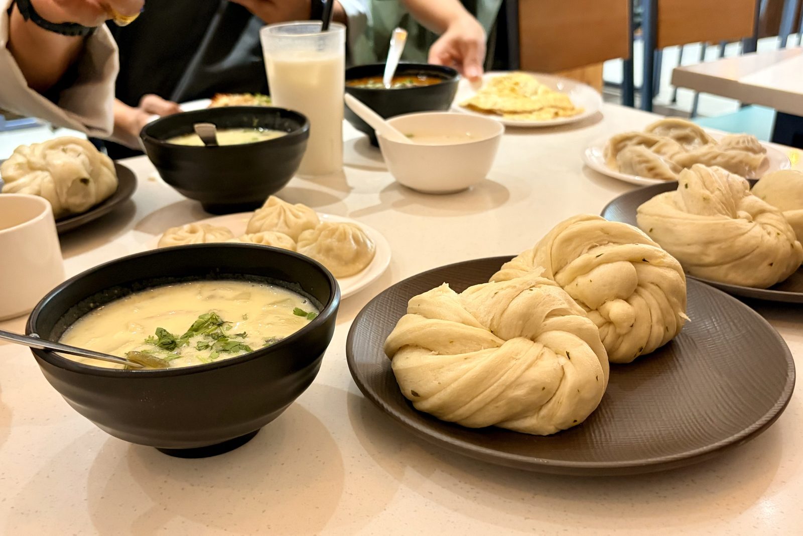 Een tafel met borden geplooide gestoomde momo's, grote gedraaide witte gestoomde broodjes, kommen romige soep bestrooid met koriander, en een hoog glas met een melkachtig drankje.