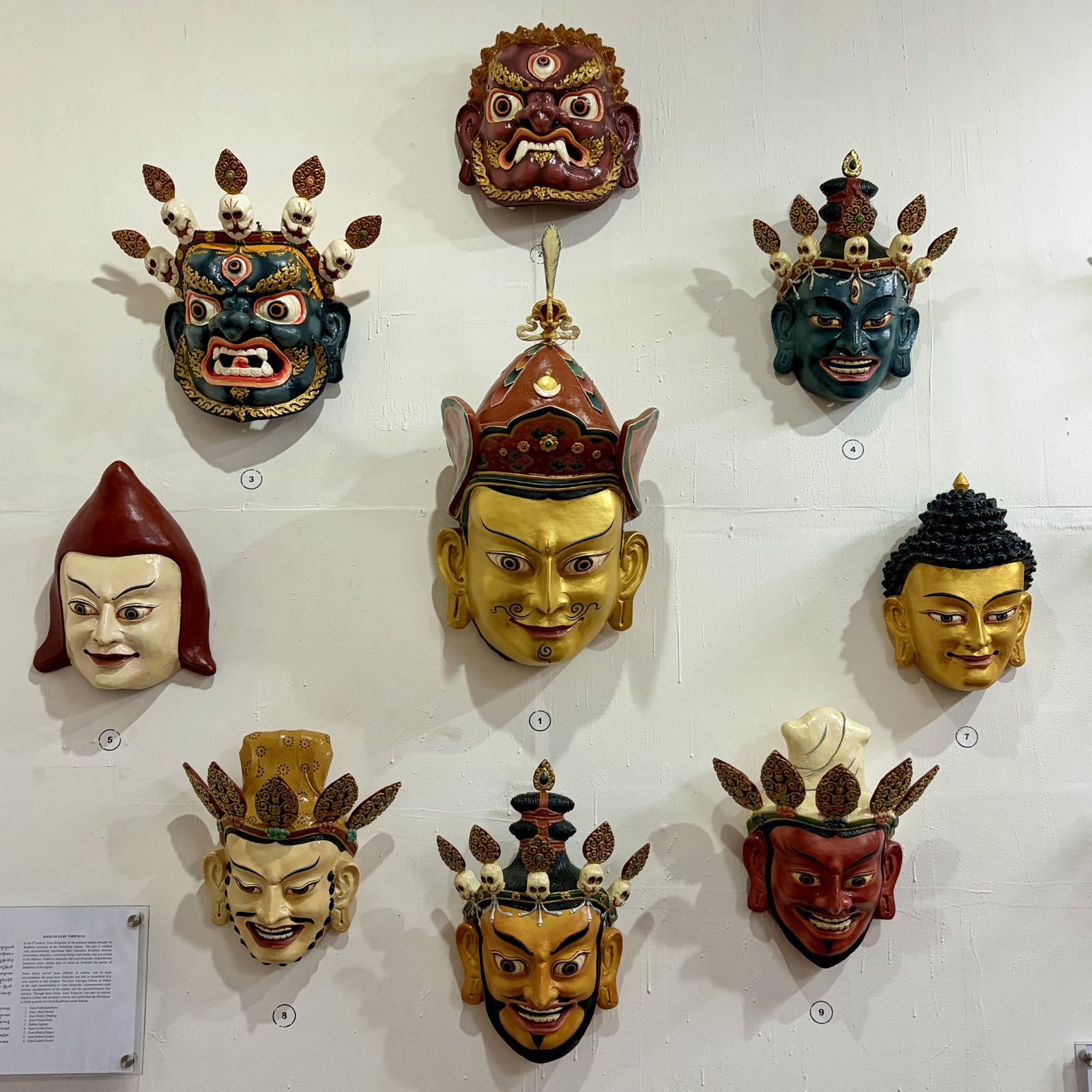 Negen beschilderde maskers aan een witte muur: in het midden een groot goudkleurig masker met een rode kroon en snor, eromheen maskers in blauw, rood, goud en wit, sommige met schedelkronen en een derde oog, met een uitlegbordje onderaan.