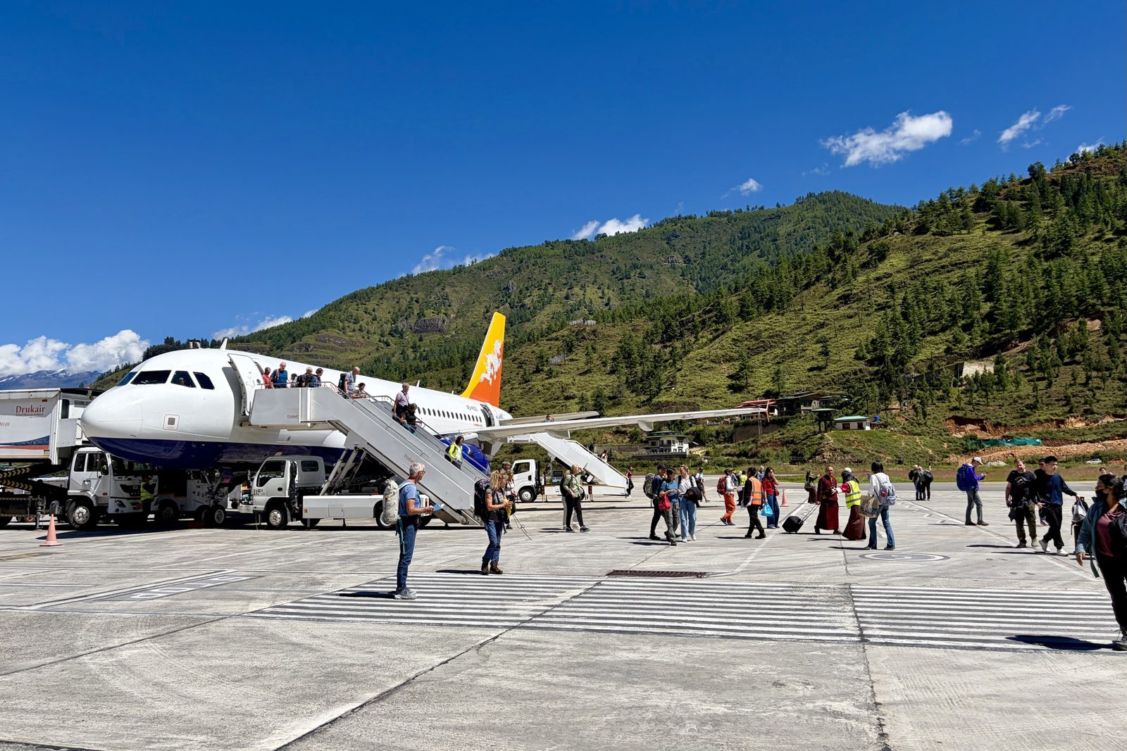 Een Drukair-toestel met een oranje draak op de staart, geparkeerd op het platform in Paro; passagiers komen de trap af en wandelen over het tarmac, met een steile, met dennen begroeide helling erachter onder een helderblauwe hemel.