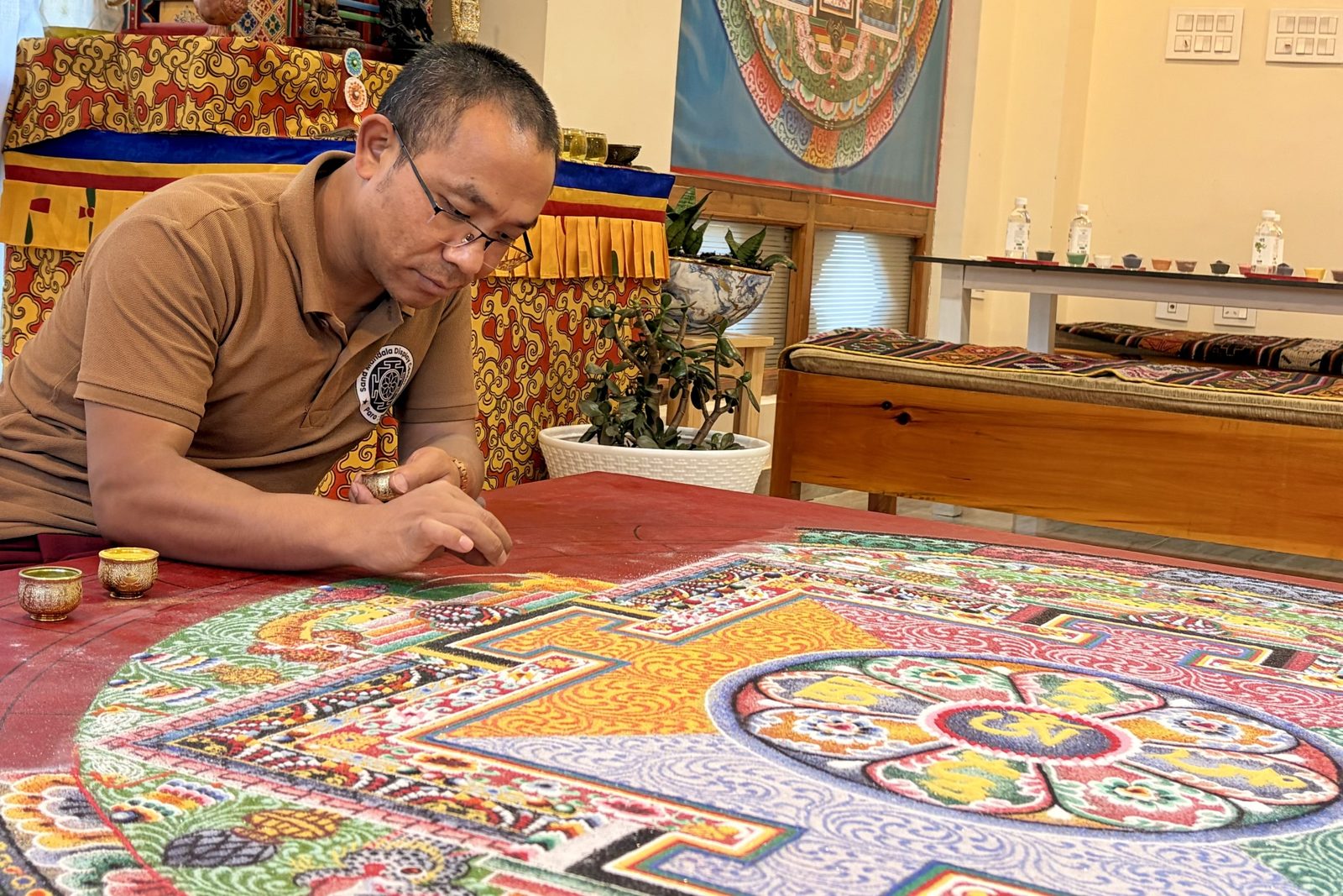 Een man met een bril buigt zich over een groot, ronde zandmandala op een rode tafel en laat met een metalen trechtertje fijn gekleurd zand vallen; de mandala heeft vierkante poorten, bloemmotieven en in het midden een lotus met gele lettergrepen.