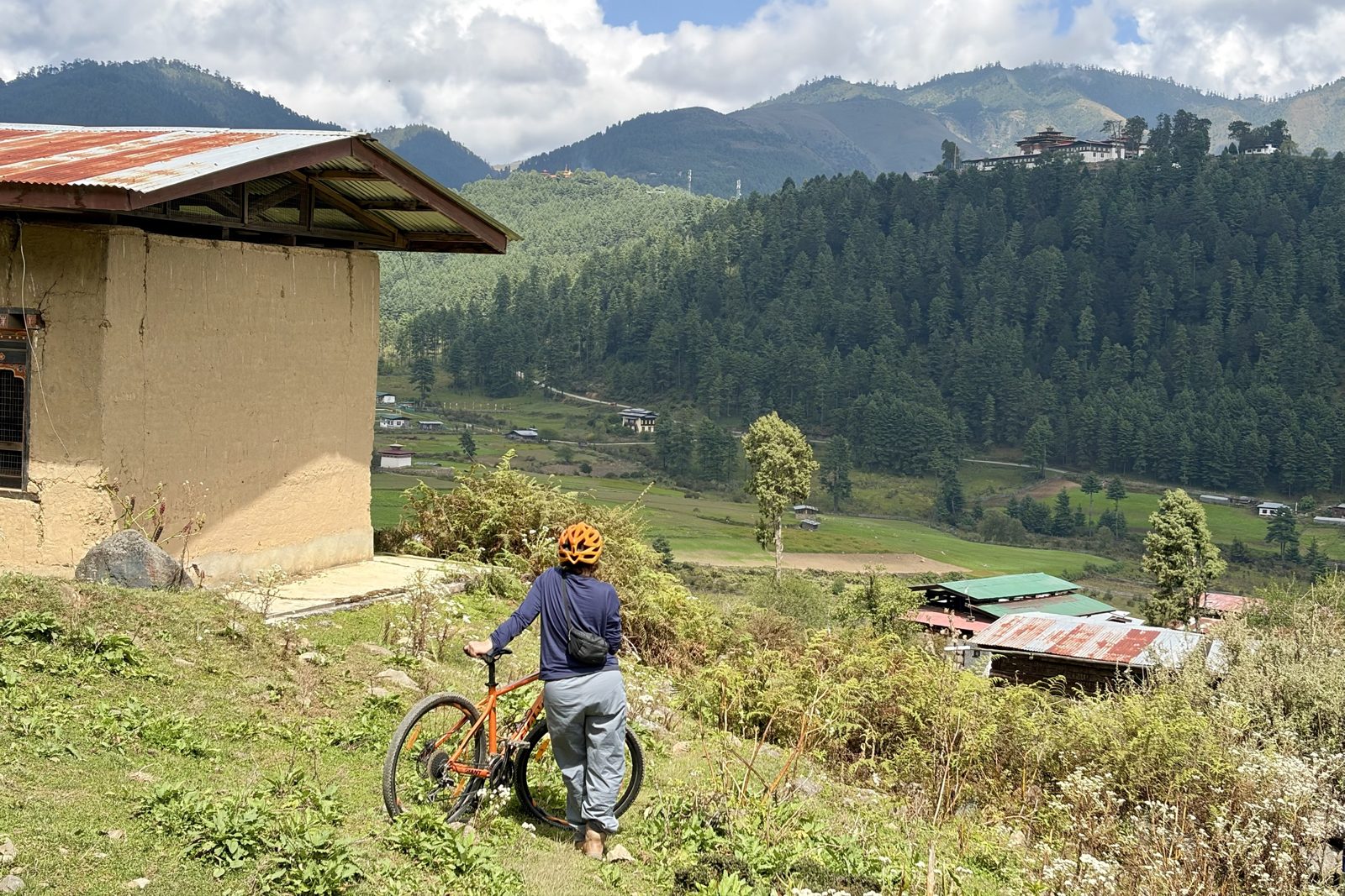 Iemand van ons met een fietshelm staat naast een oranje mountainbike bij een lemen huisje en kijkt uit over de vallei met velden, boerderijen en een beboste heuvel waarop het klooster van Gangtey ligt.