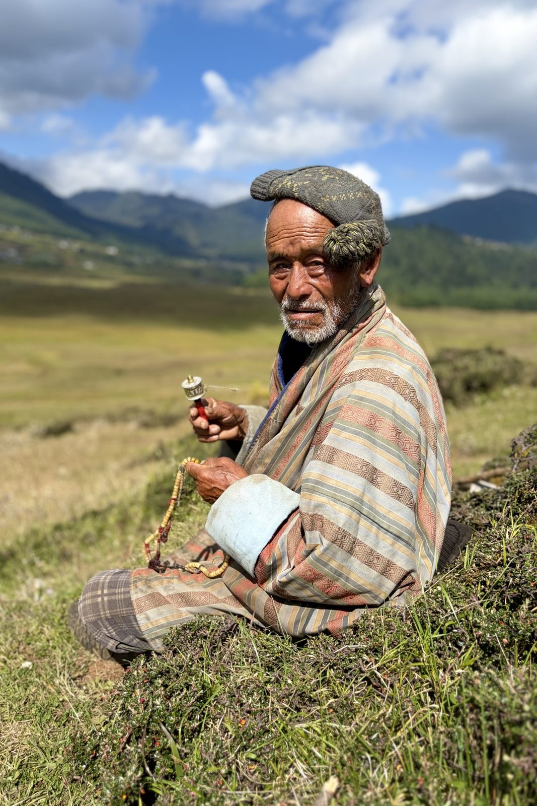 Portret van een oudere man met een witte baard en een gebreide muts, in een gestreepte gho, zittend in het gras met een kleine handgebedsmolen in de ene hand en een gebedssnoer in de andere, met de vallei vaag op de achtergrond.