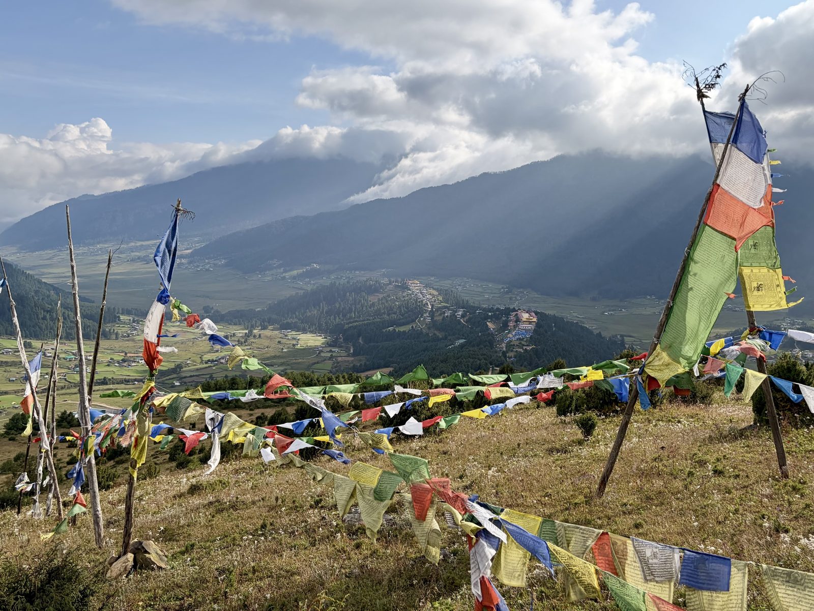 Slingers van verbleekte gebedsvlaggen in blauw, wit, rood, groen en geel tussen houten palen op een grazige helling, met de vallei van Phobjikha en zonnestralen door de wolken op de achtergrond.
