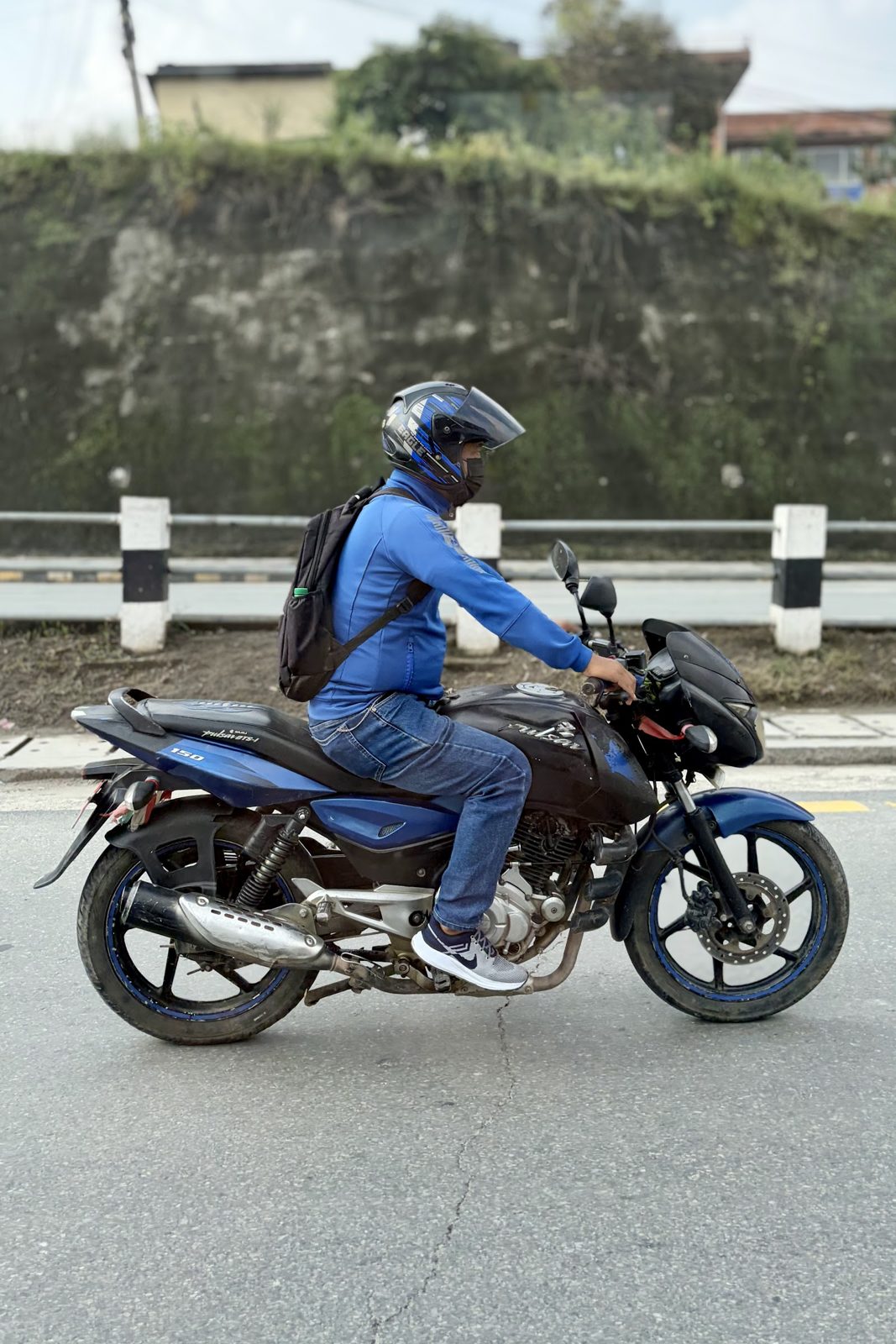 Een motorrijder met helm en blauwe jas op een blauw-zwarte motor op een asfaltweg, met een vangrail en een begroeide muur erachter.