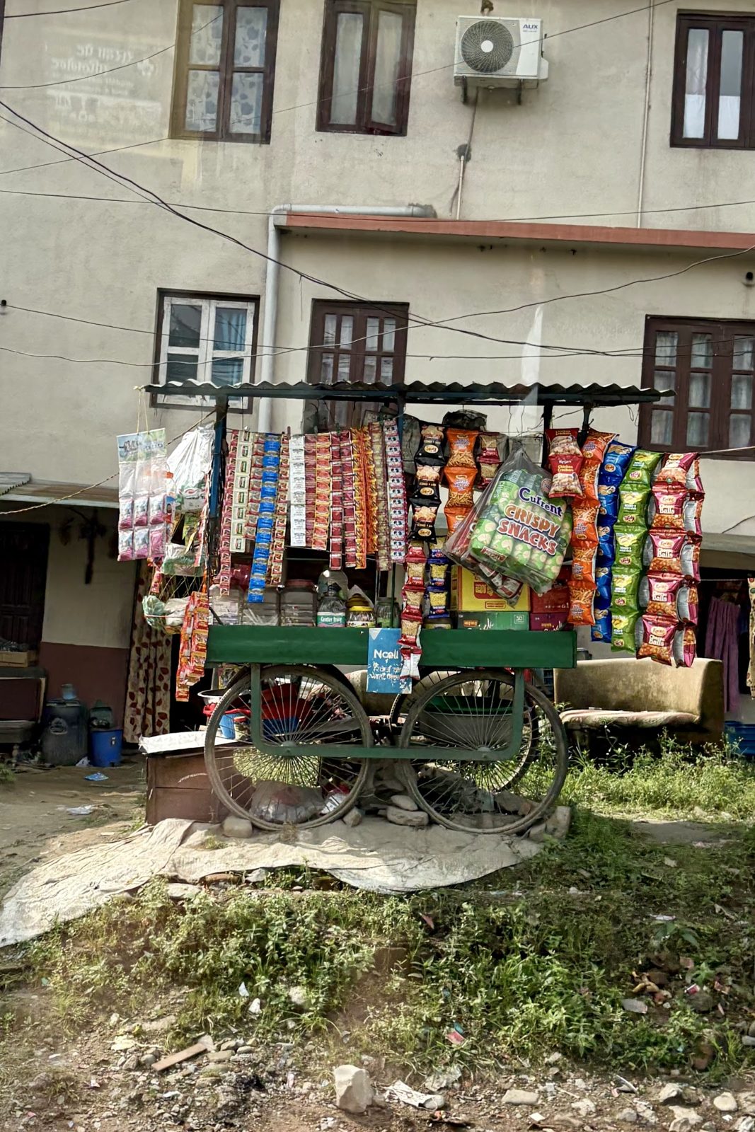 Een houten kar vol hangende zakjes snacks en kleurrijke verpakkingen langs de weg, voor een betonnen huis met balkons.