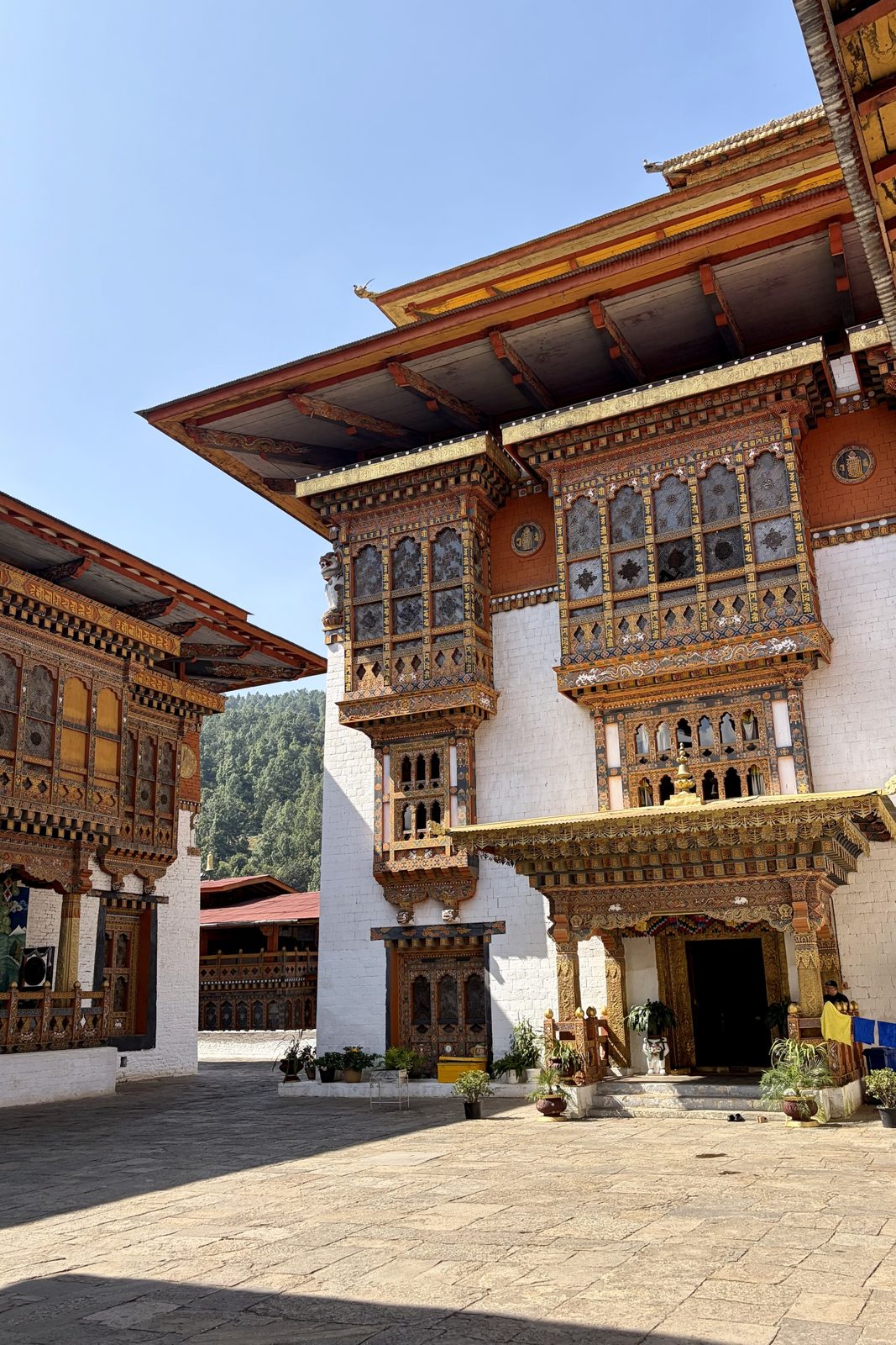 Een binnenplaats van de dzong: witte muren met grote, rijk gesneden en beschilderde houten erkers onder brede dakranden, een geplaveid plein en beboste heuvels op de achtergrond.