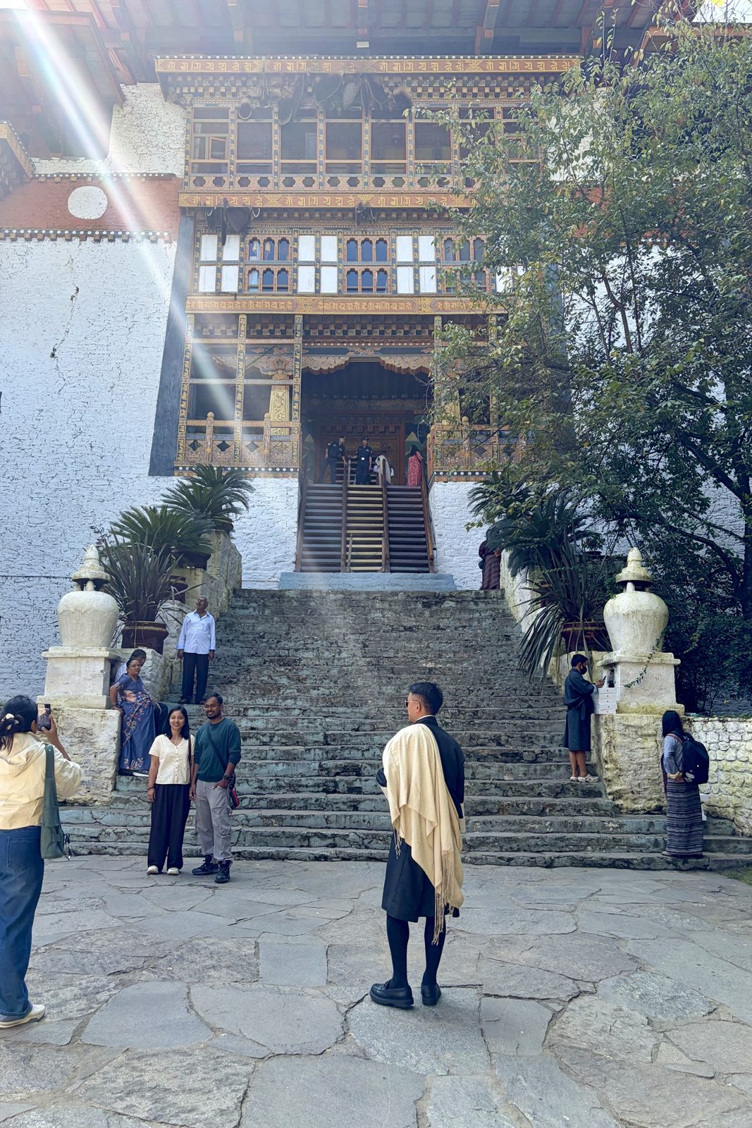 Een steile houten trap boven aan een brede stenen trap naar de ingang van de dzong, onder rijk beschilderd houtwerk; bezoekers en een man in een zwarte gho met een crèmekleurige sjaal staan op het plein ervoor.