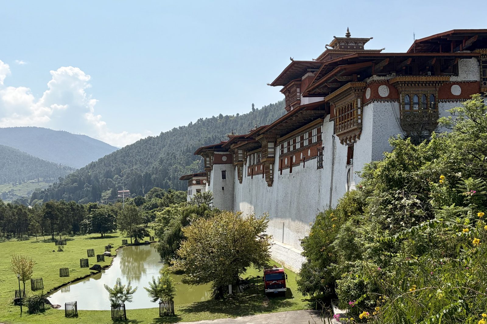 De lange witte buitenmuur van de dzong met rode banden en houten erkers, gezien vanuit de tuin: een vijver, gras, jonge boompjes in houten hekjes en een rode brandweerwagen tegen de muur, beboste heuvels erachter.