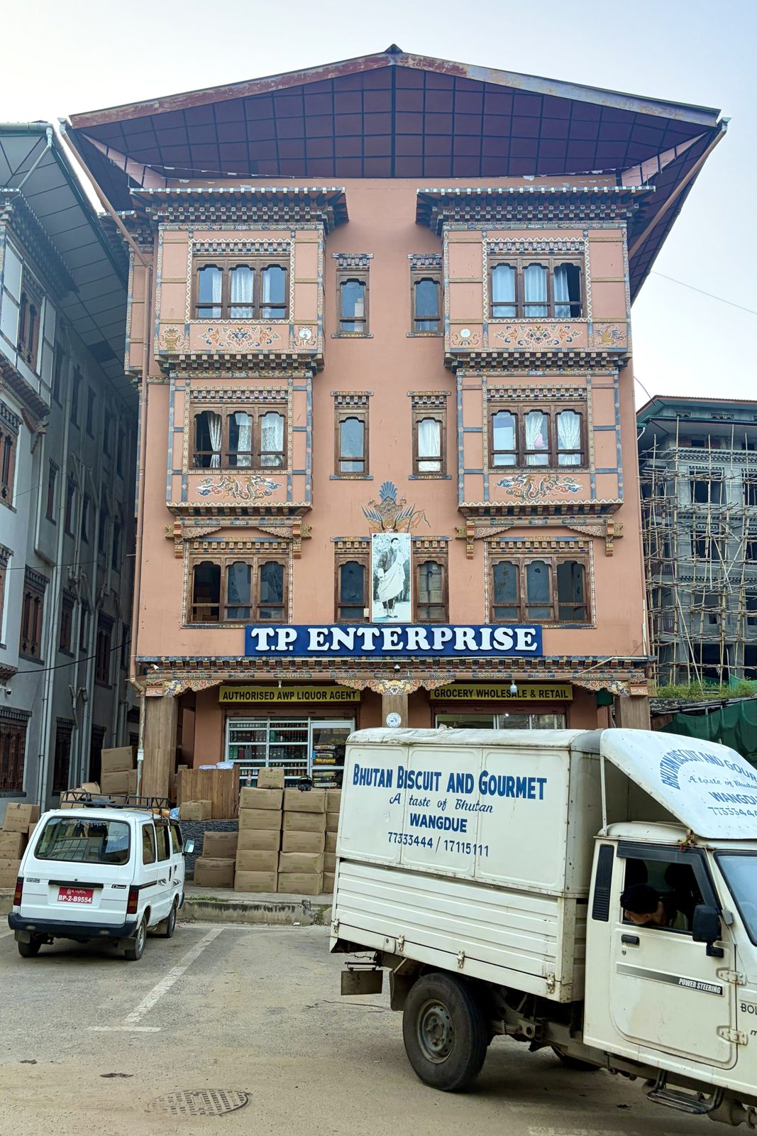 Een zalmroze gebouw met rijk beschilderde erkers, draken en een koninklijk portret op de gevel, een kruidenierszaak en drankwinkel op het gelijkvloers met dozen buiten opgestapeld, en een kleine leverwagen van Bhutan Biscuit ervoor.