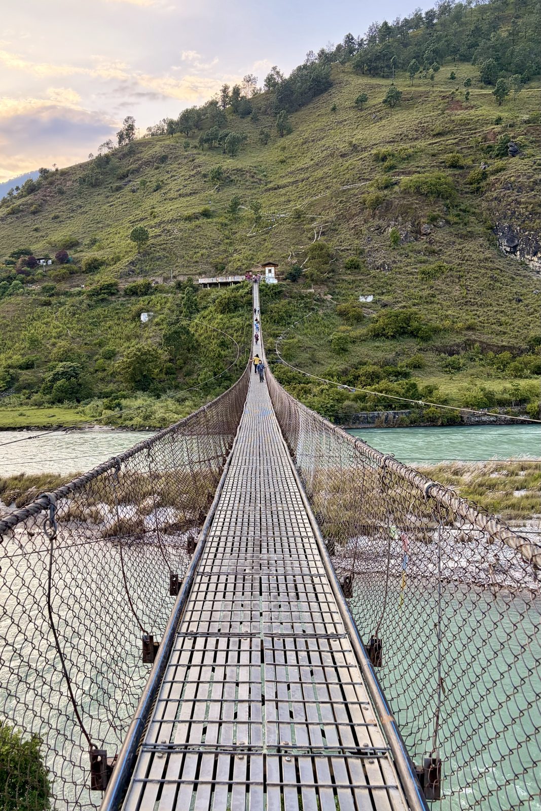 Zicht over een lange, smalle hangbrug van stalen planken met zijkanten van gaas boven een brede turquoise rivier, met mensen die oversteken richting een grazige helling, in zacht avondlicht.
