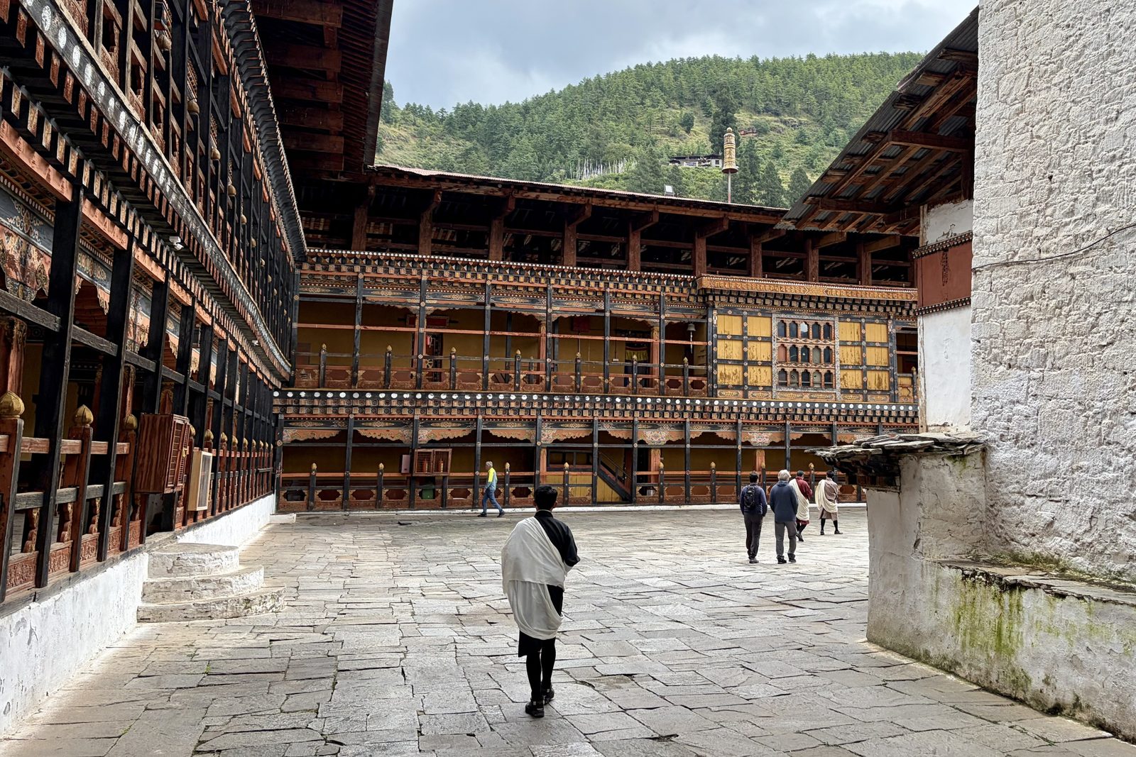 Een geplaveide binnenplaats van de dzong omringd door galerijen met rood, geel en zwart beschilderd houtwerk, met Dawa in een zwarte gho en witte kabney op de voorgrond en een paar bezoekers verderop, en een beboste helling erachter.