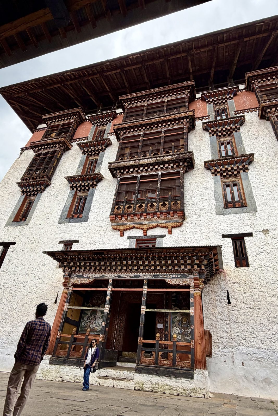 Van onderen gezien: een hoge witte gevel van de dzong met rijk gesneden en beschilderde houten vensters en erkers in bruin, rood en goud, onder een breed overstekend dak, met een bezoeker voor de ingang en iemand die omhoog kijkt.