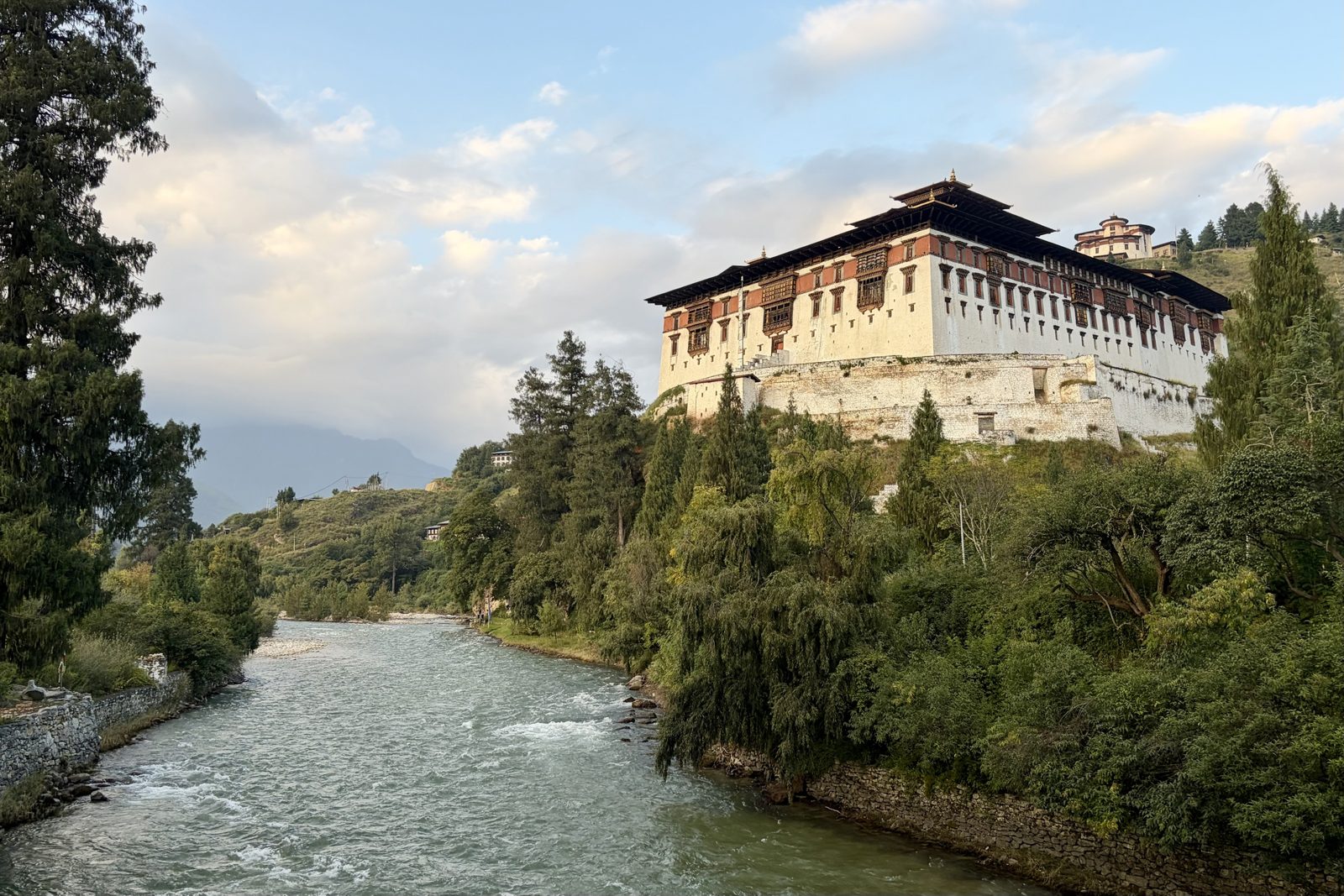 De grote witte Rinpung Dzong met rode band, houten vensters en zware daken op een heuvel boven een snelstromende rivier met bomen langs de oever, met op de heuvel erachter een rond torentje.