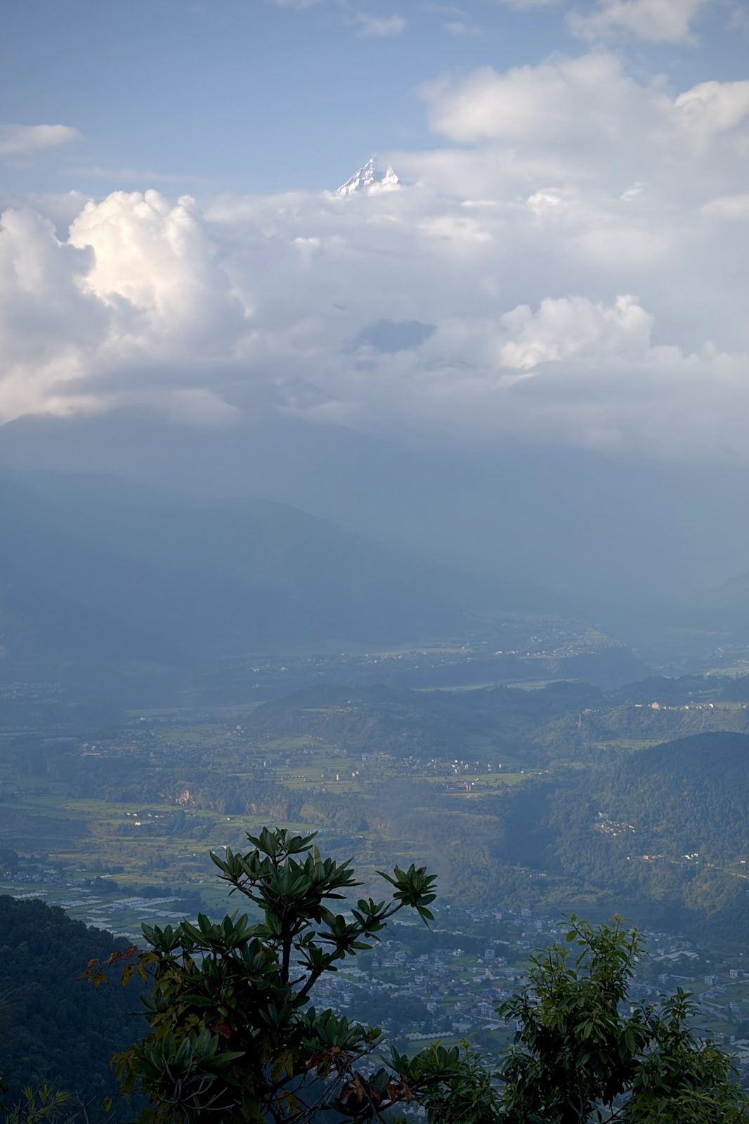 Uitzicht van op Sarangkot over de vallei van Pokhara in nevelig avondlicht, met wolken en een glimp van besneeuwde toppen.