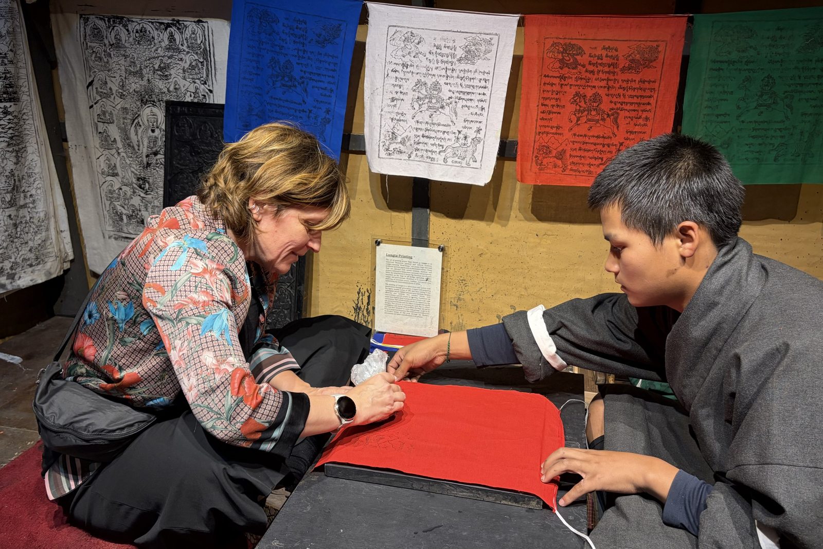 Een bezoeker wrijft rode stof over een gesneden houtblok terwijl een jonge student in een grijze gho de stof plat houdt, in een atelier vol blauwe, witte, rode en groene bedrukte gebedsvlaggen en een bordje met Lungta Printing.