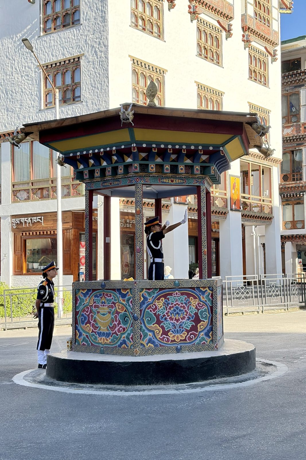 Een verkeersagent in donker uniform en met witte handschoenen regelt het verkeer met één arm in de lucht, vanuit een rijk beschilderd paviljoentje op een rotonde in Thimphu; een tweede agent staat ernaast, met witgekalkte gebouwen met gesneden, beschilderde houten ramen op de achtergrond.